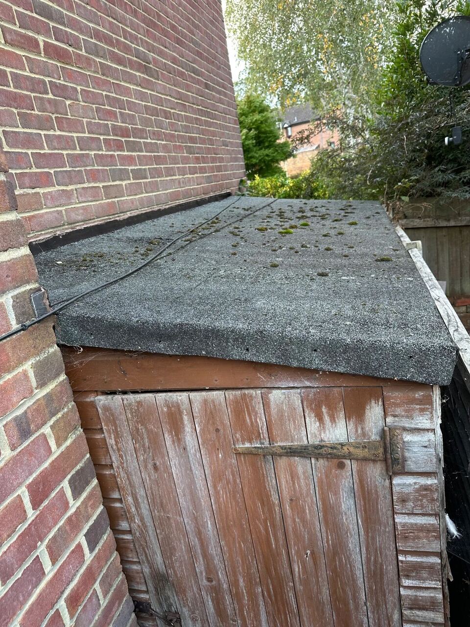 Before: weathered lean-to shed roof with moss growing on old roofing felt against a brick house wall in Yateley
